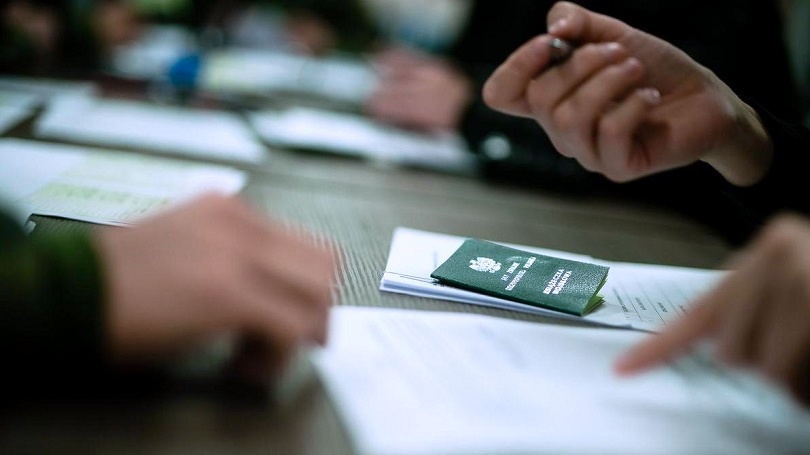 fragment stołu z dokumentami i zieloną książeczka wojskową, rozmazane zarysy trzech dłoni