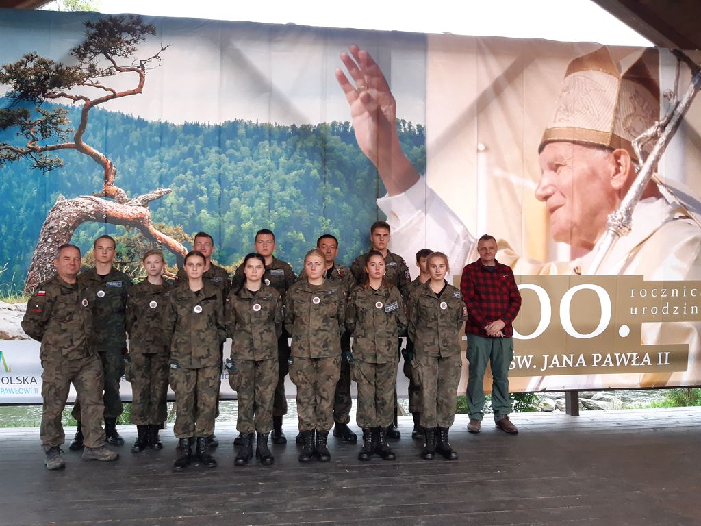 100. rocznica urodzin Jana Pawła II. Grupa uczniów w wojskowych strojach wraz z opiekunem na tle baneru promującego wydarzenie.