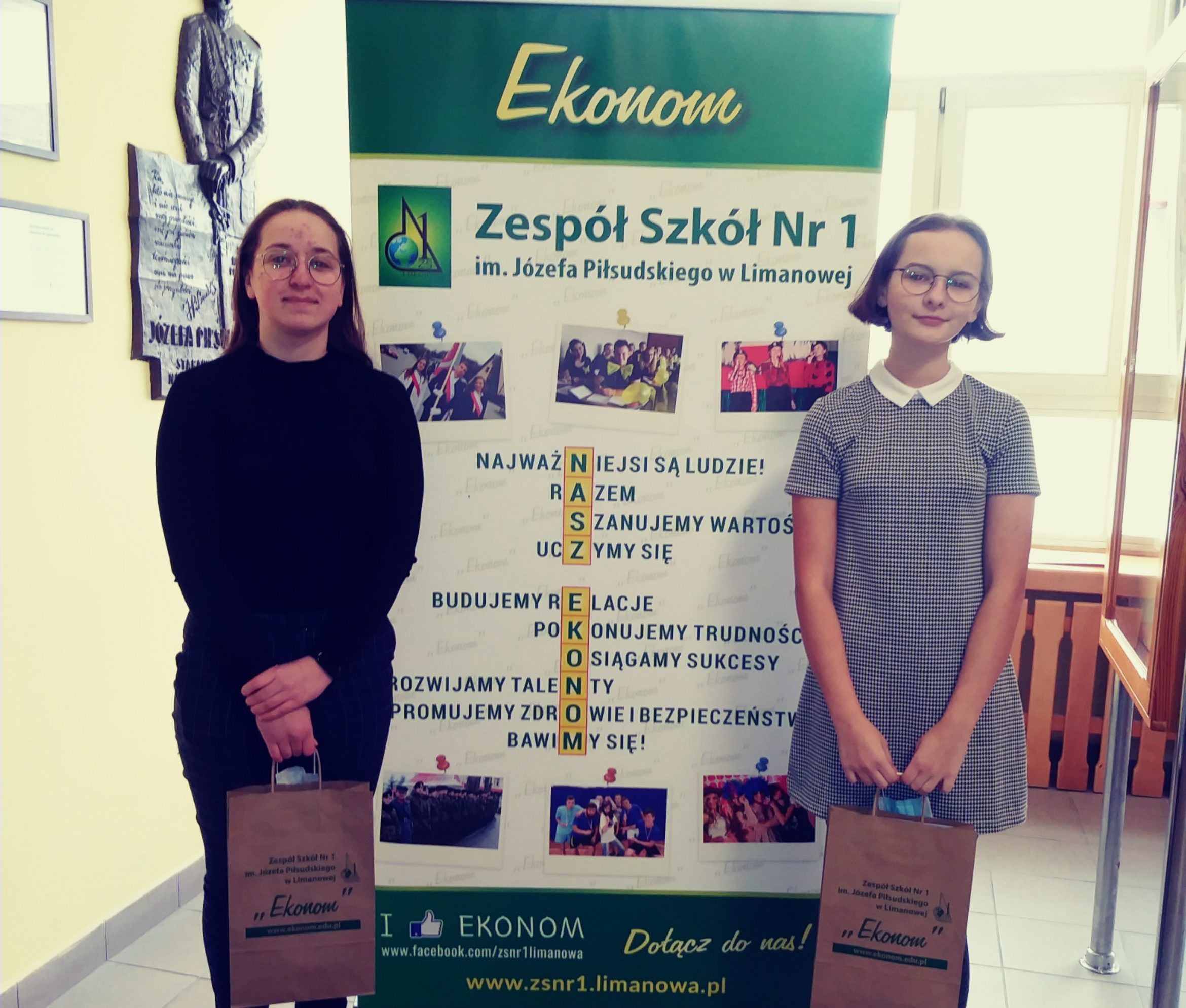 dwie dziewczyny stojące na tle rollupu szkoły trzymają torby z nagrodami.