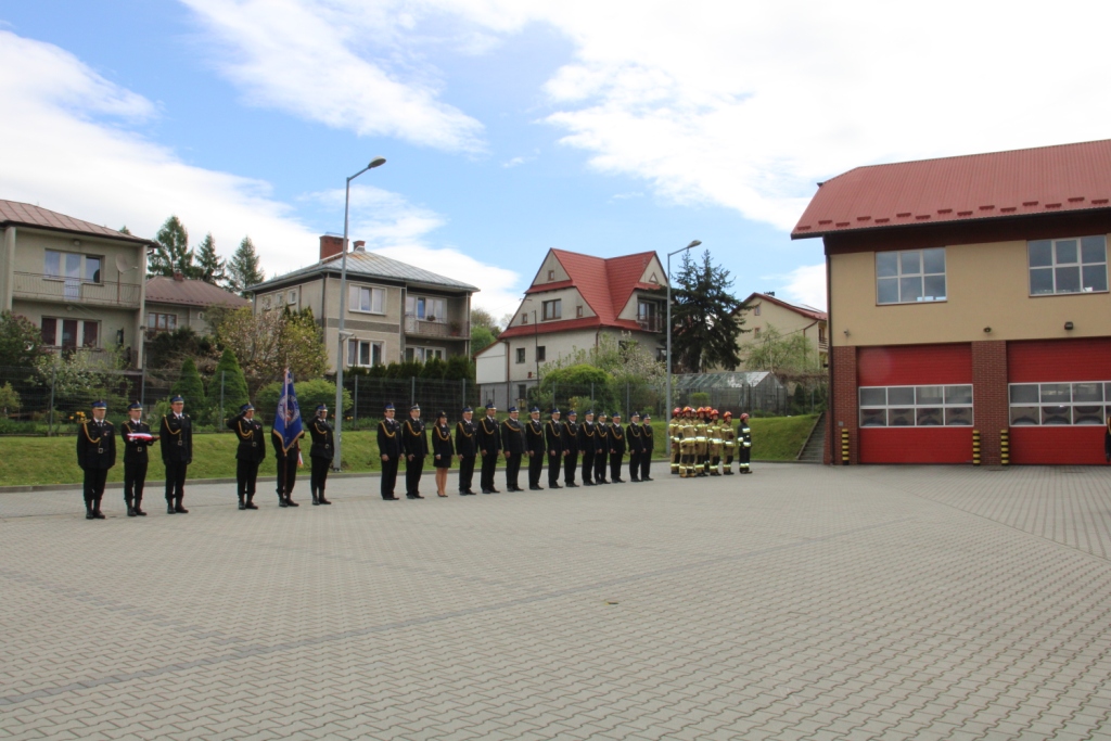 Widok na plac przed Komeda Powiatową Straży Pożarnej w Limanowej, na którym stoją ustawieni w rzedzie strażacy.