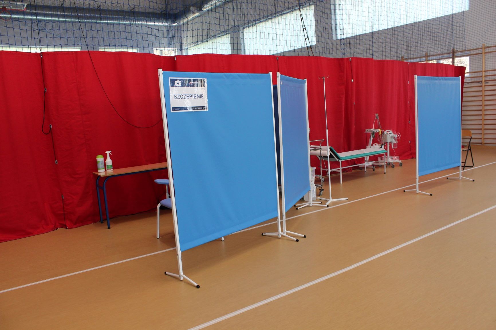 Zdjęcie części hali sportowej przygotowanej do wyknywania szczepień. Czerwone kotary i niebieskie parawany . Na zdjciu widoczne jest również łóżko dla pacjentów