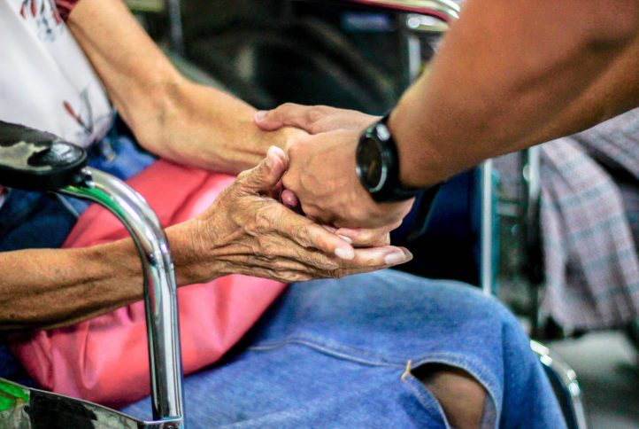 zdjęcie przedstawiające dłonie mężczyzny trzymajacego za rece starszą kobietę na wózku inwalidzkim