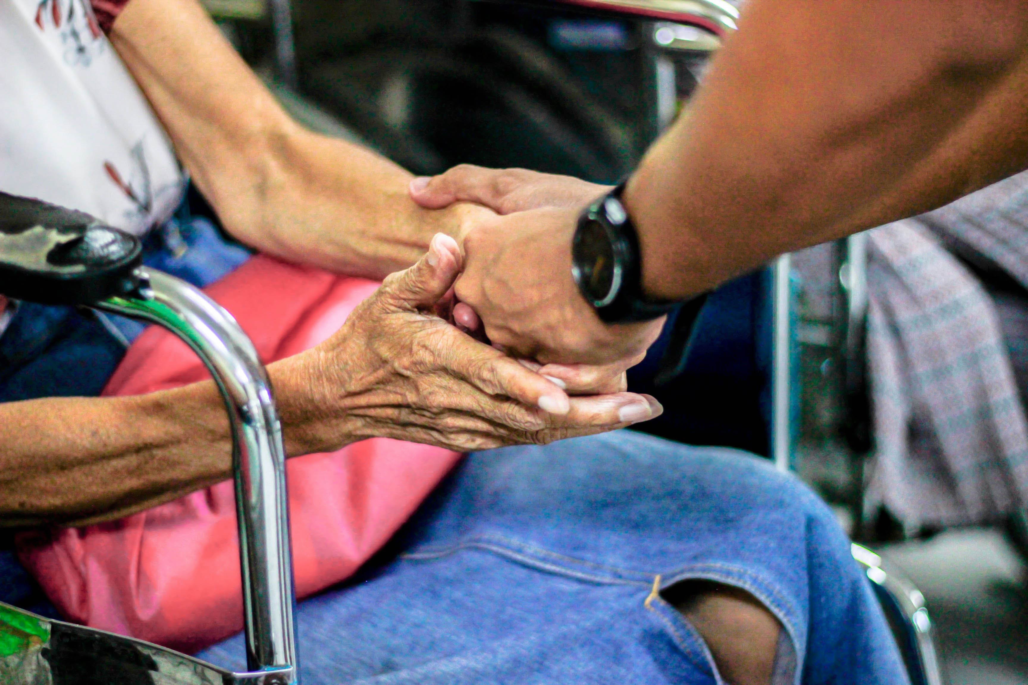 zdjęcie przedstawiające dłonie mężczyzny trzymajacego za rece starszą kobietę na wózku inwalidzkim