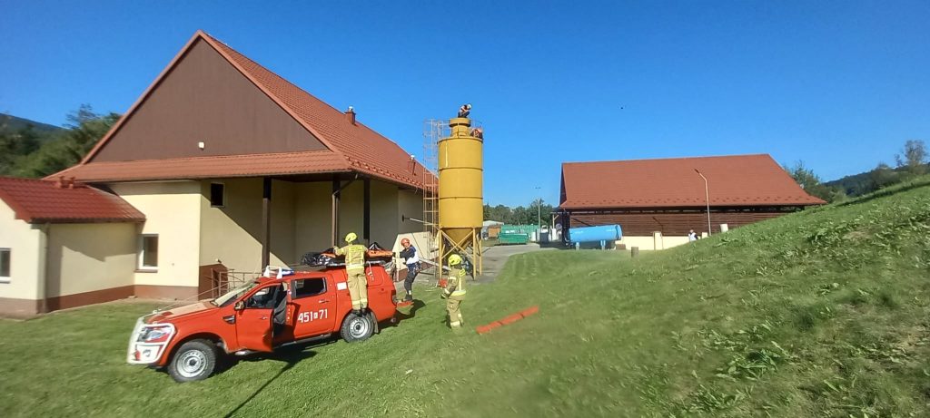 dwa budynki, żółty silos, na pierwszym planie mały samochód strażacki i trzech strażaków