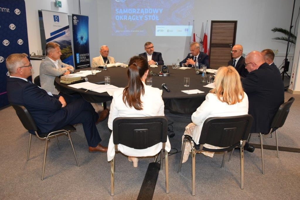 dziesięć osób siedzi przy okrągłym stole w jasnej sali, starosta Mieczysław Uryga trzyma mikrofon