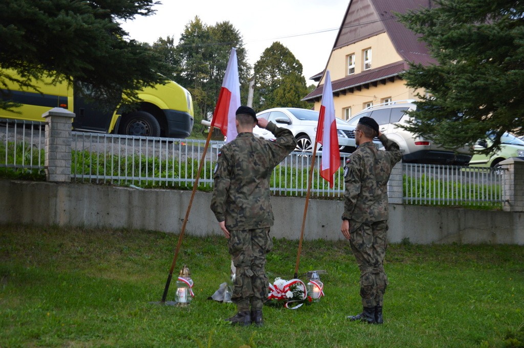 dwóch strzelców salutuje, obok flagi Polski, wiązanki i znicze