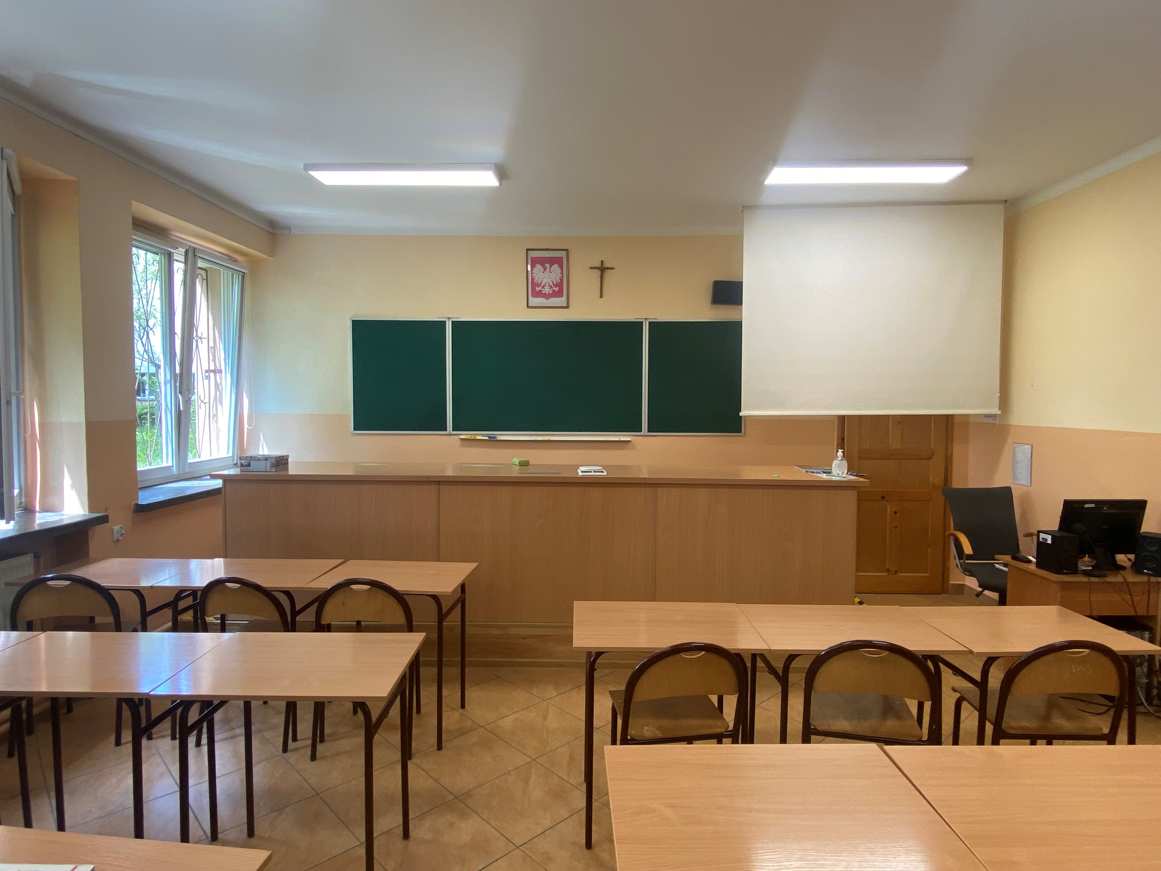 sala lekcyjna z kilkoma ławkami, duże biurko na podwyższeniu i zielona potrójna tablica