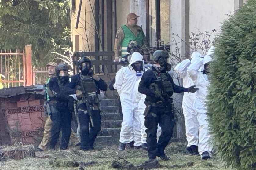 funkcjonariusze w mundurach ćwiczebnych i z bronią wyprowadzają z budynku kilka osób w białych kombinezonach i maskach gazowych