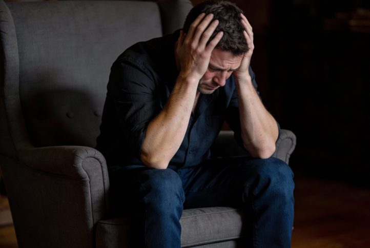 Mężczyzna siedzący na fotelu w ciemnym pomieszczeniu, trzymający się za głowę.